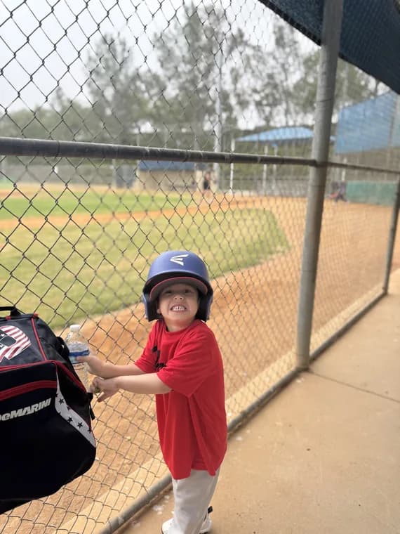 One of my boys grinning at the cage fence, helmet on, water bottle in hand.